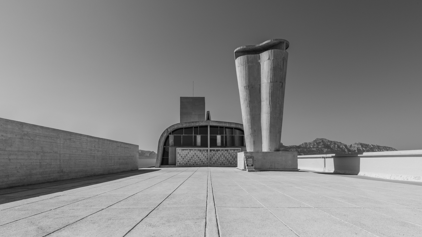 Cette photo offre une vue panoramique sur le toit de la Cité radieuse de Marseille, un célèbre bâtiment conçu par l'architecte Le Corbusier dans les années 1940. On peut y voir les différents niveaux du toit, ainsi que les bâtiments environnants et la mer Méditerranée au loin.